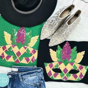 May include: A flat lay arrangement showing a black hat with a silver trim, two t-shirts with sequined mask designs, a pair of blue jeans, and silver patterned shoes. The t-shirts are green and black, with yellow, purple, and green sequin masks.