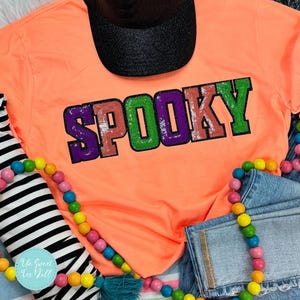 Może przedstawiać: Jasnopomarańczowa koszulka z napisem "SPOOKY" w cekinowych literach. Litery są fioletowe, zielone i pomarańczowe. Czarna czapka z daszkiem jest widoczna na górze. Kolorowe drewniane koraliki i dżinsowe szorty również znajdują się na zdjęciu.