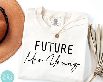 May include: Cream-coloured t-shirt with the words "FUTURE Mrs. Young" in black script. A gold necklace with a pendant rests on the shirt. A brown hat and dried plant are in the background.