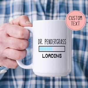 以下が含まれることがあります： 黒い文字で「DR. PENDERGRASS LOADING」と書かれた白いセラミックマグ。マグには、青いセグメントのローディングバーグラフィックがあります。「CUSTOM TEXT」という言葉が入ったピンク色の円が右上隅にあります。