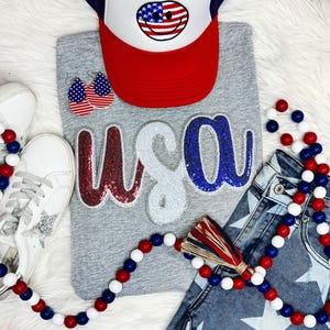 May include: A gray t-shirt with the word "USA" in red, white, and blue sequins. The shirt is paired with a red, white, and blue beaded necklace, a red, white, and blue trucker hat with a smiley face, and a pair of blue jeans with white stars.