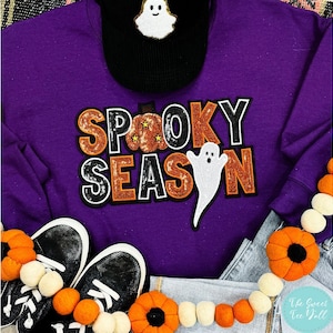 Może przedstawiać: Fioletowa bluza z napisem "Spooky Season" w pomarańczowych i białych cekinach. Czarna czapka z daszkiem z białym duchem znajduje się nad bluzą. Czarne trampki i girlanda z dyni w pierwszym planie.