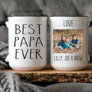 Op de afbeelding: Witte keramische mok met een zwarte handgreep en rand. De mok heeft de tekst "Best Papa Ever" gedrukt op de ene kant en een foto van drie kinderen met de tekst "Love Lilly, Joe & Drew" op de andere kant.
