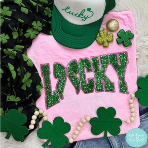 May include: A pink t-shirt with the word "LUCKY" in green glitter lettering. A green and white trucker hat with the word "lucky" is above the shirt. Decorative shamrocks and a beaded garland with shamrocks are also present.