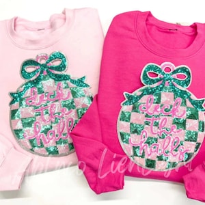 May include: Two sweatshirts, one pale pink and one cerise, each with a large sequined ornament design. The ornament features a green bow, a checkered pattern, and the phrase "Deck the Halls" in cursive.