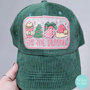 Puede incluir: Gorra de béisbol de pana verde con un parche rectangular con bordados navideños. El parche incluye una taza de café, un árbol de Navidad, un regalo y un gorro de Papá Noel, con el texto "'Tis the Season" en rosa.