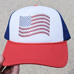 May include: A red, white, and blue trucker hat featuring a rhinestone American flag design on the front. The hat has a white front panel, a red brim, and a blue mesh back. The flag design is made of blue and red rhinestones.