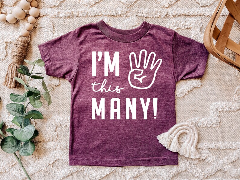 Może przedstawiać: Bordowy t-shirt z kr&oacute;tkim rękawem z białym napisem "I'm this many!" i białą ręką z uniesionymi czterema palcami.