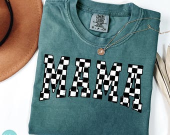 Op de afbeelding: Een teal Comfort Colors t-shirt met het woord "MAMA" in een zwart-wit geblokt patroon. Een gouden ketting met een hanger rust op de shirt. Een bruine hoed is zichtbaar op de achtergrond.