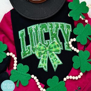 Op de afbeelding: Zwart shirt met het woord "LUCKY" in groene glitterletters en een groene geruite strik. Een zwarte hoed en een slinger van klavers en witte kralen zijn ook in de afbeelding te zien. Het shirt ligt op een roze corduroy jas.