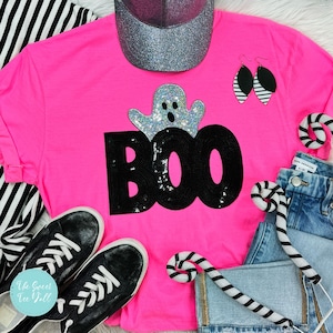 May include: A bright pink t-shirt with the word "BOO" in black sequins and a sequined ghost design. The image includes black sneakers, a silver glitter cap, black and white striped earrings, and candy cane decorations. The Sweet Tee Yall logo is visible.