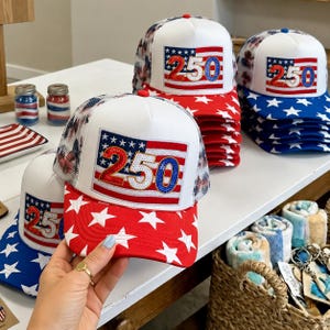 May include: A collection of patriotic trucker hats with a white crown and a red and blue star-spangled brim. Each hat features a patch with the number "250" in red and blue, set against a flag design. The hats are stacked on a white surface.