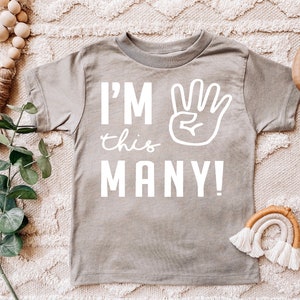 Może przedstawiać: Szara koszulka z kr&oacute;tkim rękawem z napisem "I'M this MANY!" i białym odciskiem dłoni z czterema uniesionymi palcami.