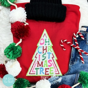 May include: A red sweatshirt with a Christmas tree design that says "Oh Christmas Tree" in colorful letters. A black knit hat sits above the sweatshirt. The image also includes denim jeans, candy canes, and a garland of pom-poms.