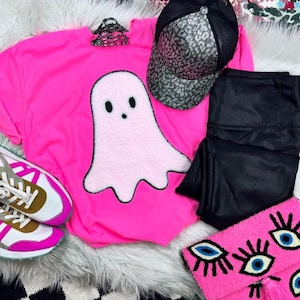 May include: A bright pink t-shirt with a light pink ghost applique, black leggings, a leopard print baseball cap, a pink clutch with eye designs, and sneakers with pink and gold accents. The items are arranged on a white faux fur surface.