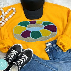 May include: Mustard yellow sweatshirt with a colourful, textured design resembling a life preserver. The design features shades of yellow, green, purple, and blue. A black baseball cap and black trainers are also visible.