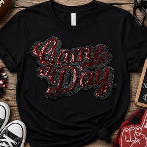 Puede incluir: Una camiseta negra de manga corta con el texto Game Day en lentejuelas rojas y un contorno de purpurina negra. La prenda tiene cuello redondo y está confeccionada en tela negra lisa. El diseño está centrado en la parte delantera.