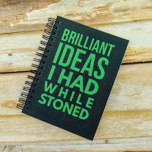 May include: A black spiral notebook with the text "BRILLIANT IDEAS I HAD WHILE STONED" printed in green.