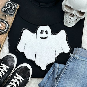 Puede incluir: Sudadera negra con un aplique de fantasma blanco sonriente y un contorno de lentejuelas negras. La imagen también incluye zapatillas negras, vaqueros azules y una calavera.