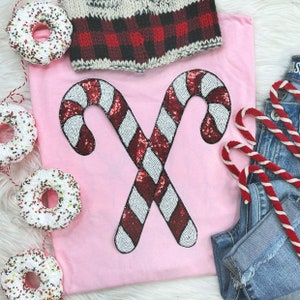 May include: A light pink t-shirt featuring two crossed candy canes embellished with red and white sequins. The image also includes a red and black plaid knit hat, denim jeans, and decorative donuts with sprinkles.