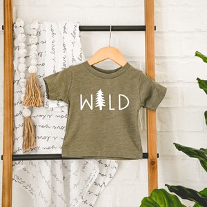 Może przedstawiać: Zielona koszulka z kr&oacute;tkim rękawem z napisem "WILD" w białych literach i małym białym drzewem sosny.