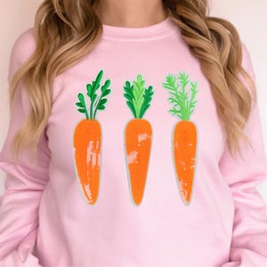 Puede incluir: Una sudadera rosa claro con tres zanahorias naranjas bordadas con hojas verdes. Las zanahorias están dispuestas verticalmente y tienen una textura de cuentas. La sudadera es de estilo cuello redondo y manga larga.