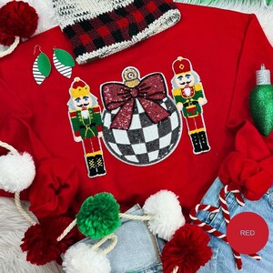 Puede incluir: Un suéter rojo festivo con un gran diseño de adorno adornado con un patrón de cuadros y un lazo de lentejuelas. Dos figuras de cascanueces flanquean el adorno. Los accesorios incluyen un gorro a cuadros, aretes verdes y guirnalda de pompones.