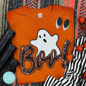 May include: Orange Halloween-themed t-shirt with a sequined ghost and the word "Boo!". The shirt is paired with black and white striped earrings and decorative pumpkins, perfect for the spooky season.
