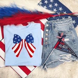 May include: A light blue t-shirt with a patriotic bow design, paired with denim shorts and a small flag that says "Red, White & Boom." The image also includes red, white, and blue decorative elements.