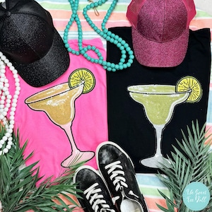 May include: Two black t-shirts with a sequined margarita design. One shirt is pink and the other is black. The shirts are laid out on a striped beach towel with a black mesh cap, a pink glitter trucker hat, a turquoise beaded necklace, and white beaded necklace, and black sneakers.