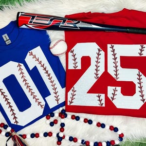 May include: Two baseball jerseys, one blue with the number "01" and one red with the number "25". Both numbers are white with red stitching and are decorated with rhinestones. A baseball bat and a baseball are also in the image.