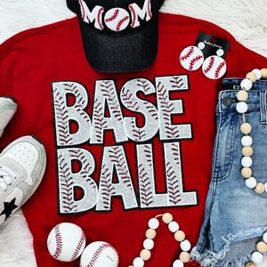 May include: Red t-shirt with the word "BASEBALL" in white rhinestones. The shirt is decorated with baseball stitching and has a red, white, and blue baseball cap on top. There are also white and brown beaded necklaces and baseball earrings.
