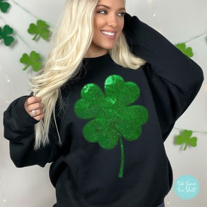 Puede incluir: Sudadera negra con un gran diseño de trébol de lentejuelas verdes brillantes. El trébol está centrado en la parte delantera. El logotipo de The Sweet Tee Doll es visible en la esquina inferior derecha.