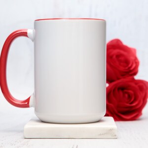 May include: White ceramic mug with a red rim and handle. The mug is sitting on a white marble coaster. There are red roses in the background.