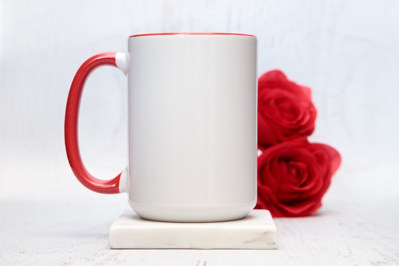 May include: White ceramic mug with a red rim and handle. The mug is sitting on a white marble coaster. There are red roses in the background.
