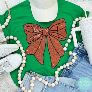May include: A green t-shirt with a large sparkly orange bow on the front. The shirt is laying on a white surface with a white and brown beaded garland and a pair of white sneakers.