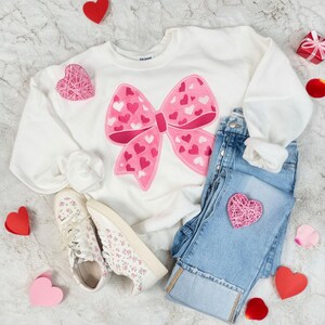 Op de afbeelding: Een witte sweatshirt met een groot roze strikdesign bedekt met kleine hartjes, gecombineerd met een lichtblauwe jeans en bloemen sneakers. Een roze hart patch en verschillende papieren hartjes maken de Valentijnsdag outfit compleet.