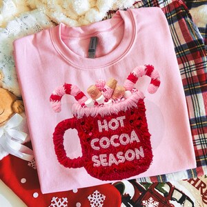 Puede incluir: Sudadera rosa claro con un diseño bordado en rojo de una taza de chocolate caliente. La taza está llena de malvaviscos y bastones de caramelo y tiene la inscripción "HOT COCOA SEASON".
