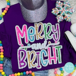 May include: A purple sweatshirt with the words "Merry and Bright" in colorful sequined letters. A silver glitter baseball cap and a snowflake ornament are also visible. The sweatshirt is paired with denim jeans and colorful beads.