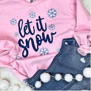 May include: A pale pink sweatshirt featuring the words "let it snow" in navy blue script, decorated with sparkly blue snowflakes. The sweatshirt is styled with blue denim shorts and a garland of white pom-poms.