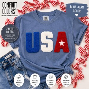 May include: A blue jean coloured t-shirt featuring the word "USA" in textured letters. The "U" is blue, the "S" is white, and the "A" is red with a white star. The shirt has a relaxed fit and is garment-dyed.