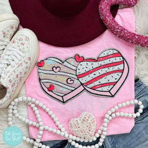 Op de afbeelding: Roze T-shirt met twee pailletten hartvormige ontwerpen met rode en witte strepen en kleine hartjes. De afbeelding bevat bloemen sneakers, een bordeauxrode hoed en een kralenketting. Er is ook een decoratief hart aanwezig.