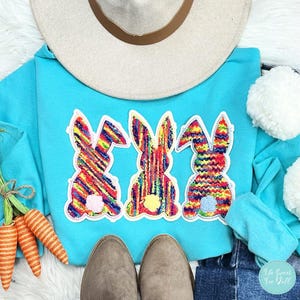 May include: A turquoise hoodie with three colourful bunny appliqués. The bunnies have a rainbow pattern and are outlined in white. A beige hat and a pair of grey boots are also visible. The image has an Easter theme.