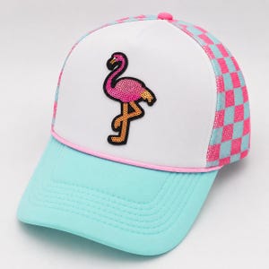 Puede incluir: Gorra de camionero con un panel frontal blanco con un flamenco rosa con lentejuelas. La visera es azul claro y los laterales de malla tienen un estampado de tablero de ajedrez rosa y azul claro. El sombrero tiene un botón rosa en la parte superior y un ribete rosa.