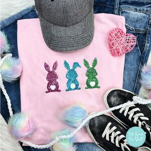 May include: A light pink t-shirt with three glittery bunny designs in purple, blue, and green, each with a white heart. A grey baseball cap, denim jeans, and black trainers with white laces are also in the image.