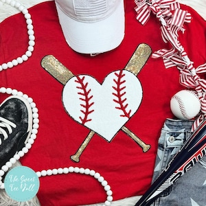 May include: Red t-shirt with a white heart-shaped baseball and two gold baseball bats crossed behind it. The shirt is surrounded by white beads, a white baseball cap, a pair of blue jeans, and a red and white striped ribbon.