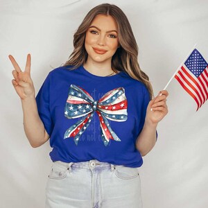 May include: A royal blue t-shirt featuring a watercolor bow design in red, white, and blue, with stars and stripes. The person is holding a small American flag and making a peace sign.