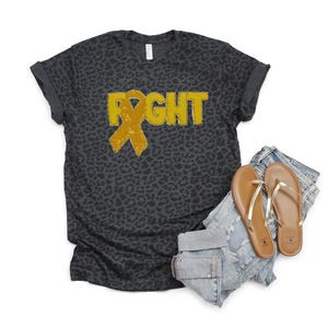 May include: Dark gray leopard print t-shirt with the word "FIGHT" in yellow letters and a yellow ribbon graphic. The shirt is paired with light blue denim shorts and brown sandals with gold straps.