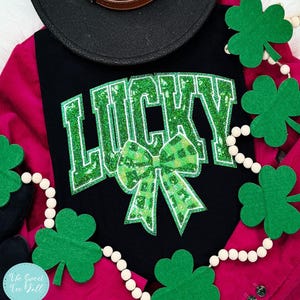 Puede incluir: Camiseta negra con la palabra "LUCKY" en letras verdes brillantes y un lazo verde a cuadros. Una gorra negra y una guirnalda de tréboles y cuentas blancas también están en la imagen. La camiseta está sobre una chaqueta de pana rosa.