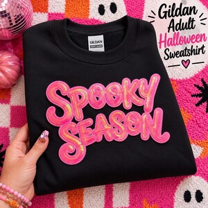 Op de afbeelding: Een zwart Gildan Heavy Blend sweatshirt met de tekst "SPOOKY SEASON" in grote, felroze, iriserende paillettenletters op de borst. Het kledingstuk heeft een ronde hals, lange mouwen en een wit merklabel aan de binnenzijde van de kraag.